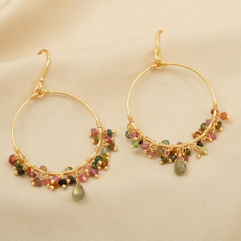 Puede incluir: Un par de pendientes de aro dorados. Cada pendiente presenta un dise&ntilde;o de alambre dorado con racimos de peque&ntilde;as cuentas de colores en tonos rosa, verde y marr&oacute;n. Una cuenta en forma de l&aacute;grima cuelga de la parte inferior de cada pendiente.