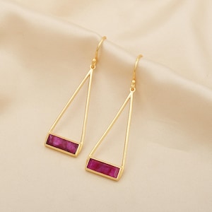 以下が含まれることがあります： 幾何学的なデザインのゴールドカラーのイヤリングのペア。各イヤリングは、ベースに長方形のマゼンタ色の石が付いた三角形のフレームを備えています。イヤリングは、クリーム色のサテンのような生地に表示されています。
