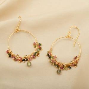 Puede incluir: Un par de pendientes de aro dorados. Cada pendiente est&aacute; adornado con un grupo de peque&ntilde;as piedras preciosas de colores en tonos rosa, verde y negro. Una sola piedra preciosa en forma de l&aacute;grima cuelga de la parte inferior de cada pendiente.