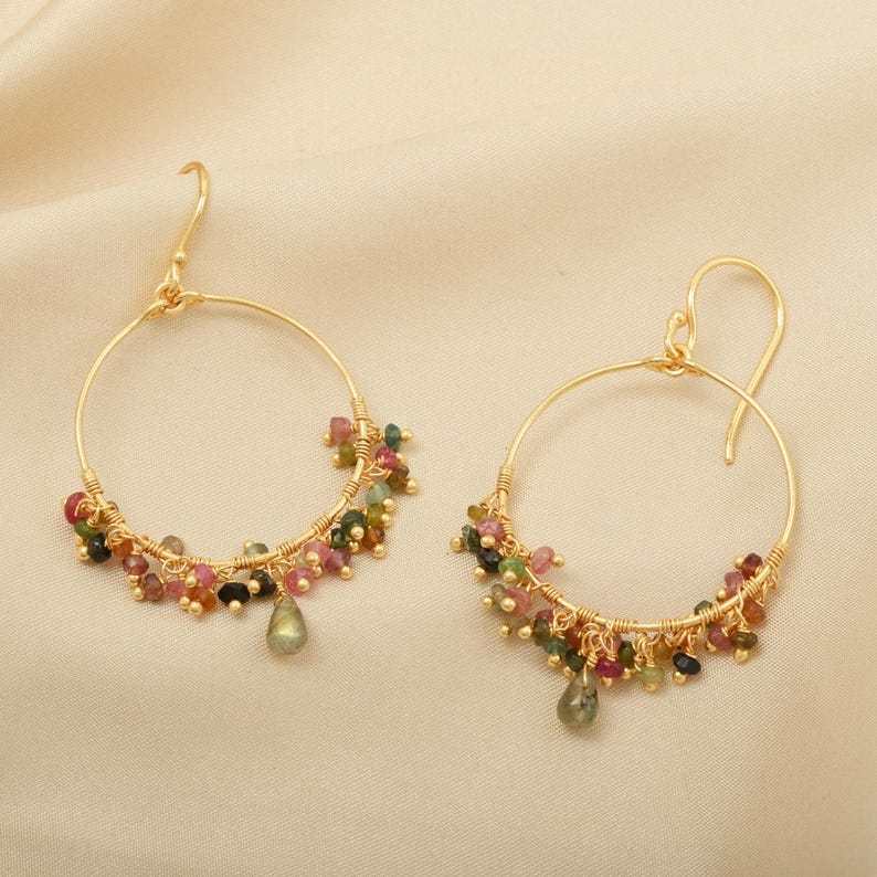 Puede incluir: Un par de pendientes de aro dorados. Cada pendiente est&aacute; adornado con un grupo de peque&ntilde;as piedras preciosas de colores en tonos rosa, verde y negro. Una sola piedra preciosa en forma de l&aacute;grima cuelga de la parte inferior de cada pendiente.