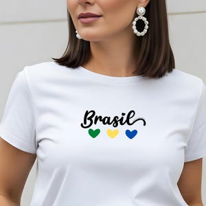 May include: White t-shirt with the word "Brasil" in black script, accompanied by green, yellow, and blue heart symbols. The shirt is made of a soft material and is suitable for casual wear.