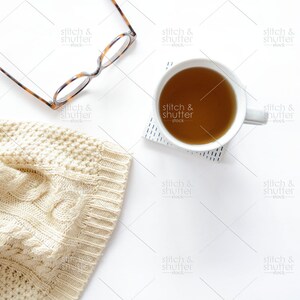 May include: A white ceramic mug filled with brown tea sits on a white surface with a black and white patterned coaster. A pair of tortoiseshell glasses and a cream-colored knitted sweater are also on the surface.