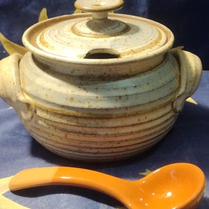 May include: A white ceramic soup tureen with a lid and two handles. The tureen has a brown and white speckled glaze and a brown and white striped pattern. A matching orange ladle is included.