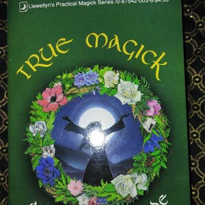 May include: A green book cover with the title "True Magick: A Beginner's Guide" by Amber K. The cover features a wreath of flowers surrounding a silhouette of a person with arms raised against a blue sky with a full moon.