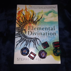 May include: A book titled "Elemental Divination: A Dice Oracle" by Stephen Ball. The cover features a sun and moon with flames and clouds. There are six dice in front of the book, including a black die, a white die, a blue die, a purple die, and two green dice.