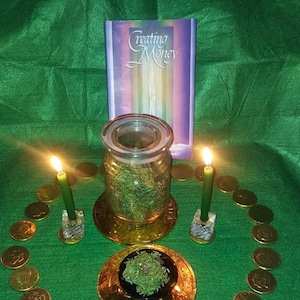 May include: A green table cloth with a circle of gold coins, two green candles, a glass jar filled with green herbs, and a book titled "Creating Money".