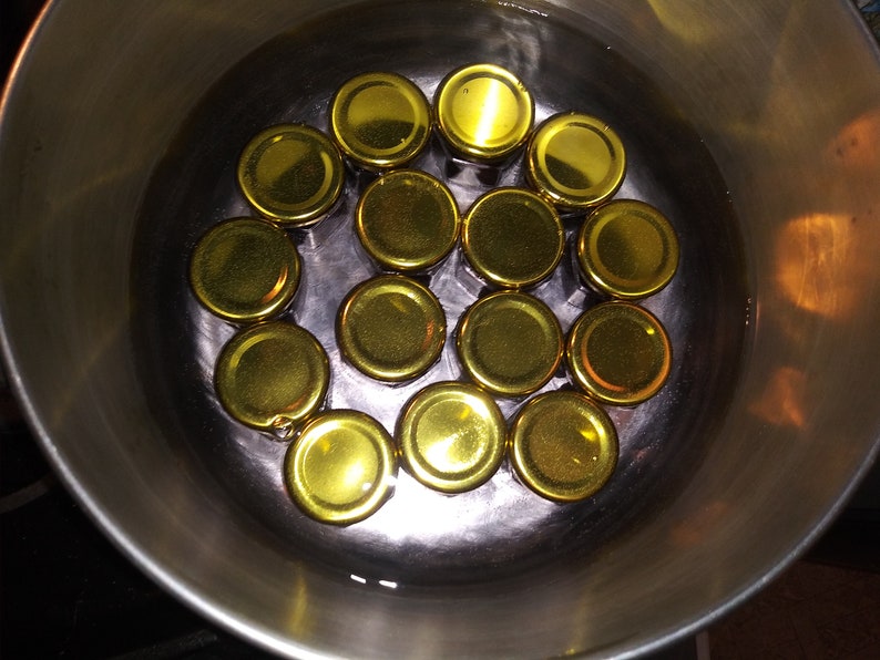 May include: A close-up shot of a stainless steel pot filled with water and several golden-colored jar lids. The lids are arranged in a circular pattern, suggesting they are being sterilized or prepared for canning.