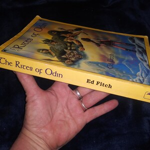 Może przedstawiać: Ż&oacute;łta książka w miękkiej oprawie zatytułowana "The Rites of Odin" autorstwa Ed Fitch. Okładka przedstawia kolorową ilustrację grupy ludzi w fantastycznym otoczeniu.