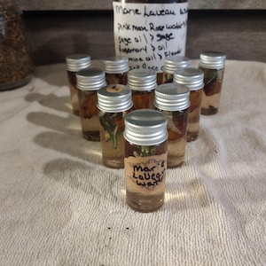 May include: A collection of small glass bottles filled with liquid and herbs, topped with silver lids. The bottles are arranged on a white cloth. Some bottles have labels that read "Marie Laveau Water". A handwritten note is visible in the background.