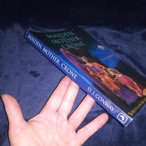 May include: A book titled "Maiden, Mother, Crone: The Mysteries of the Triple Goddess" by D.J. Conway. The cover features a painting of three women in flowing robes, set against a dark blue night sky with a crescent moon.