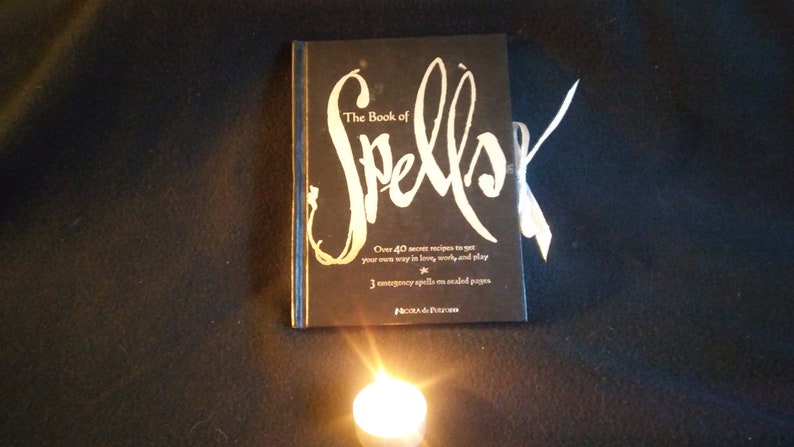 May include: A black book with a silver title that reads "The Book of Spells". The book has a white ribbon bookmark and is sitting on a black surface with a lit candle in front of it.
