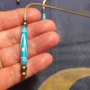 May include: A pair of earrings with a brass-colored curved wire and a turquoise bead with white accents. The earrings have small brass-colored beads at the top and bottom of the turquoise bead.