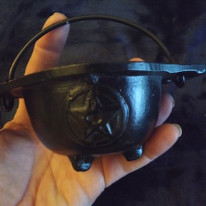 May include: A small, black cast iron cauldron with a handle and three small feet. The cauldron features a pentagram design on the front. The handle is arched, and the cauldron is held in a hand.