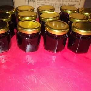 May include: A collection of small, hexagonal glass jars filled with a dark, reddish-purple preserve. Each jar has a gold-colored lid. The jars are arranged on a bright pink surface, suggesting a homemade or artisanal product, possibly jam or jelly.