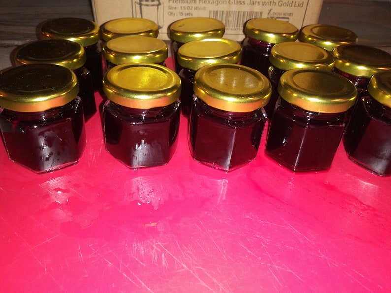 May include: A collection of small, hexagonal glass jars filled with a dark, reddish-purple preserve. Each jar has a gold-colored lid. The jars are arranged on a bright pink surface, suggesting a homemade or artisanal product, possibly jam or jelly.