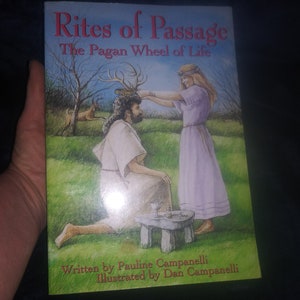 May include: Book cover with an illustration of a woman placing a crown of antlers on a man's head. The book title is "Rites of Passage: The Pagan Wheel of Life". The book is written by Pauline Campanelli and illustrated by Dan Campanelli.