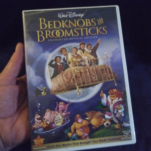 May include: DVD case for the Walt Disney movie Bedknobs and Broomsticks, featuring a scene of the characters flying on a bed with broomsticks. The case is blue with a gold border and features the movie title in white text. The text "Enchanted Musical Edition" is also included in white text. The case also features a picture of the characters from the movie, including the main characters, the bed, and the broomsticks.