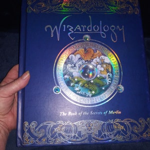 May include: A blue hardback book titled "Wizardology" with gold foil detailing and a circular illustration. The book, "The Book of the Secrets of Merlin," has a detailed cover design with a holographic effect.