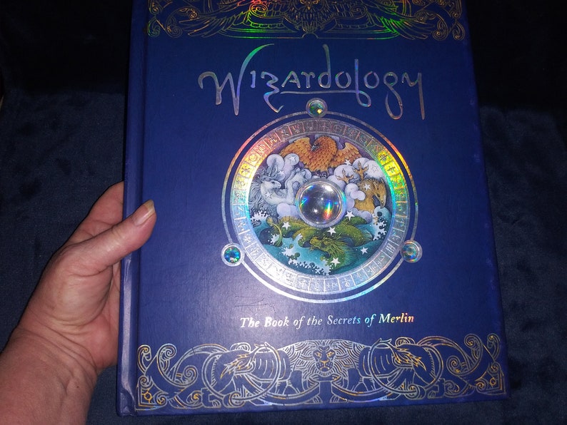 May include: A blue hardback book titled "Wizardology" with gold foil detailing and a circular illustration. The book, "The Book of the Secrets of Merlin," has a detailed cover design with a holographic effect.