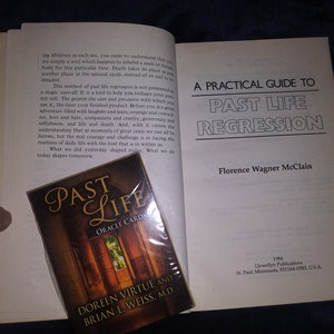 Puede incluir: Un libro blanco con el t&iacute;tulo "A Practical Guide to Past Life Regression" de Florence Wagner McClain. El libro est&aacute; abierto para revelar una p&aacute;gina con texto sobre la regresi&oacute;n de vidas pasadas. Una baraja de cartas de or&aacute;culo titulada "Past Life Oracle Cards" de Doreen Virtue y Brian L. Weiss, M.D. es visible en primer plano.