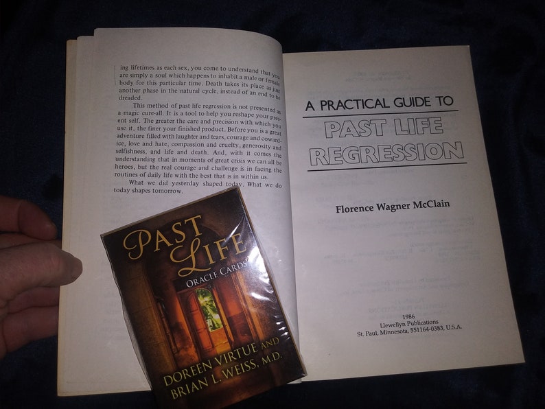 Puede incluir: Un libro blanco con el t&iacute;tulo "A Practical Guide to Past Life Regression" de Florence Wagner McClain. El libro est&aacute; abierto para revelar una p&aacute;gina con texto sobre la regresi&oacute;n de vidas pasadas. Una baraja de cartas de or&aacute;culo titulada "Past Life Oracle Cards" de Doreen Virtue y Brian L. Weiss, M.D. es visible en primer plano.