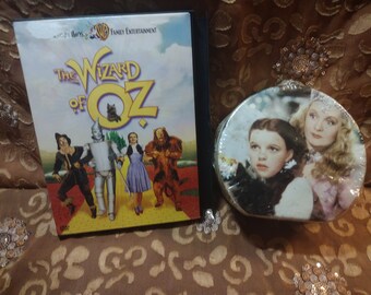 May include: A DVD case for the movie "The Wizard of Oz" featuring a yellow brick road and the main characters Dorothy, the Scarecrow, the Tin Man, and the Cowardly Lion. A round tin container with a picture of Dorothy and Glinda the Good Witch is also shown.
