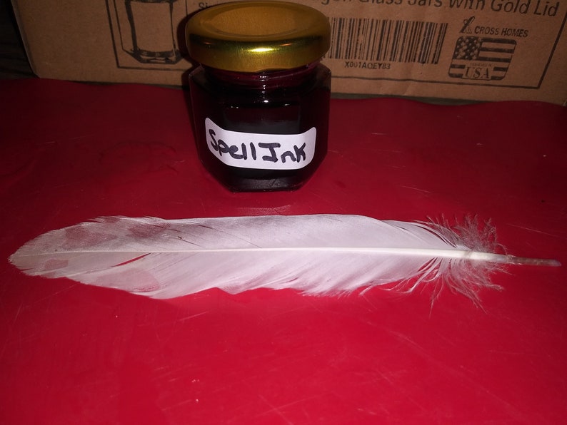 May include: A small glass jar of dark red "Spell Ink" with a gold lid, next to a white feather. The jar has a white label with the words "Spell Ink" in black. The items are on a red surface.