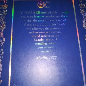 May include: A dark blue book cover with gold and iridescent foil accents. The cover features ornate designs and text that reads, "IF YOU ARE unshakable in your desire to learn wizardology...Merlin, 1577."