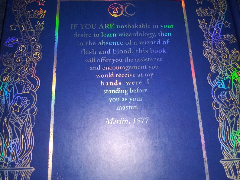 May include: A dark blue book cover with gold and iridescent foil accents. The cover features ornate designs and text that reads, "IF YOU ARE unshakable in your desire to learn wizardology...Merlin, 1577."