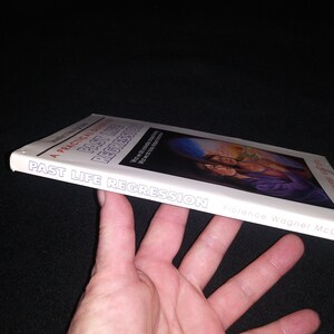 May include: A white book titled "Past Life Regression: A Practical Guide" by Florence Wagner McClain. The book cover features a painting of two people embracing in a field with a sunset in the background.