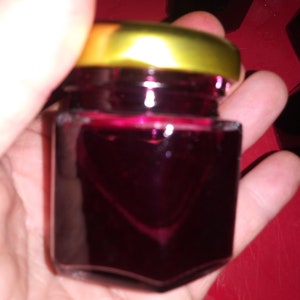 May include: A small, square glass jar filled with a deep red liquid, possibly jam or jelly. The jar has a gold-colored lid. The image is taken close-up, with the jar held in a person's hand. Other jars are visible in the background.