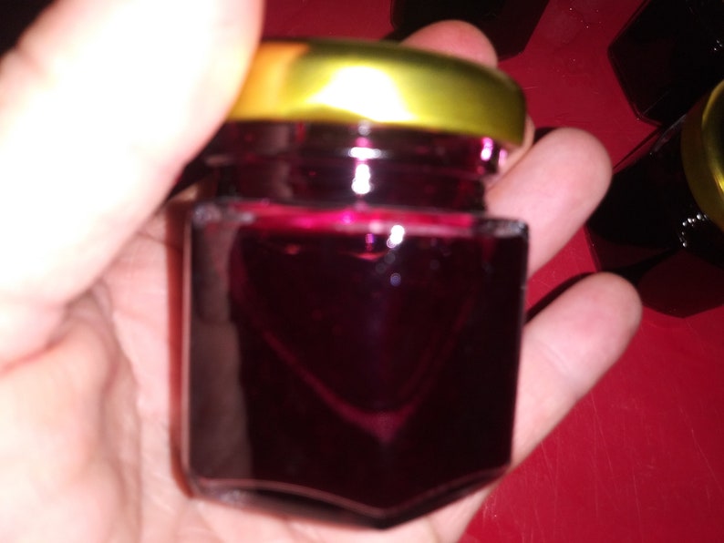 May include: A small, square glass jar filled with a deep red liquid, possibly jam or jelly. The jar has a gold-colored lid. The image is taken close-up, with the jar held in a person's hand. Other jars are visible in the background.