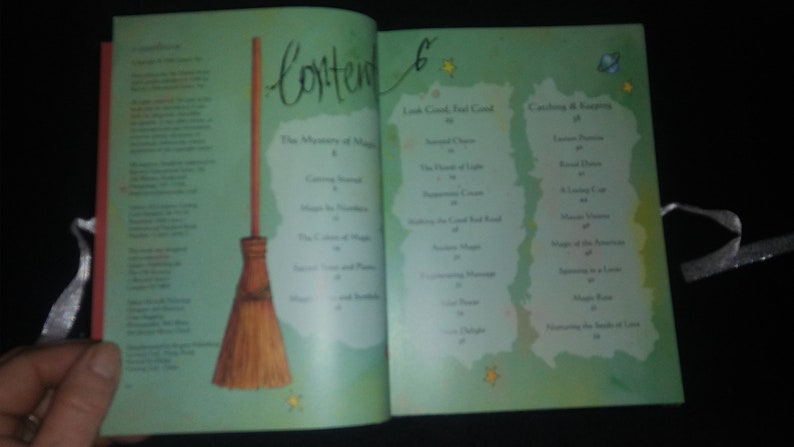 May include: An open book with a green cover and a table of contents. The book features a title called "The Mysteries of Magic" and a list of chapters including "Catching Rainbows" and "The Legend of the Red Road". A broom is illustrated on the left page.