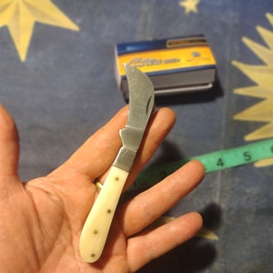 May include: A small, white-handled pocket knife with a curved blade. The blade is made of stainless steel and has a slight curve to it. The handle is made of white plastic and has a few small, gold-colored dots on it.