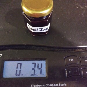 May include: A small jar of dark liquid labeled "Spell Ink" sits on an electronic scale, weighing 3.4 ounces. The jar has a gold lid and a white label with black text. The scale is black.
