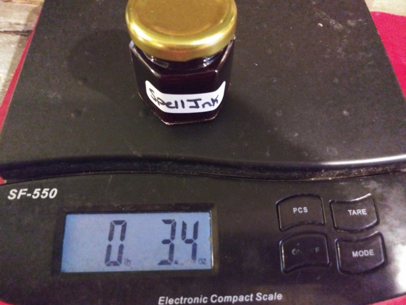May include: A small jar of dark liquid labeled "Spell Ink" sits on an electronic scale, weighing 3.4 ounces. The jar has a gold lid and a white label with black text. The scale is black.