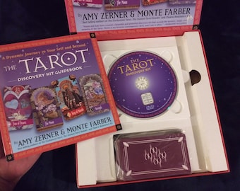 May include: A purple and white tarot card deck discovery kit with a guidebook, DVD, and cards. The guidebook has a red cover with the title "The Tarot Discovery Kit Guidebook" and features images of tarot cards. The DVD is purple with the title "The Tarot Discovery Kit". The cards are in a red box with a purple and white design.