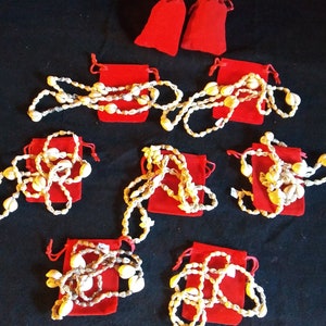 May include: Several shell necklaces displayed on a black surface. Each necklace is made of small, cream-colored shells strung together. The necklaces are presented on red velvet pouches, with two additional pouches visible.