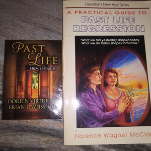 Puede incluir: Un libro titulado "A Practical Guide to Past Life Regression" de Florence Wagner McClain, parte de la serie New Age de Llewellyn. La portada del libro presenta una pintura de dos mujeres mirando por una ventana una puesta de sol sobre el oc&eacute;ano. Junto al libro hay una baraja de cartas de or&aacute;culo titulada "Past Life Oracle Cards" de Doreen Virtue y Brian L. Weiss, M.D.