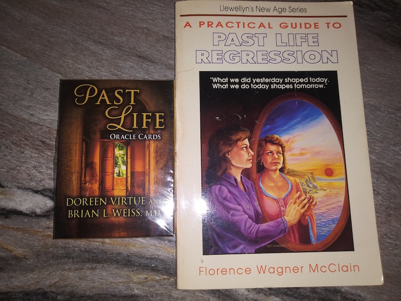 Puede incluir: Un libro titulado "A Practical Guide to Past Life Regression" de Florence Wagner McClain, parte de la serie New Age de Llewellyn. La portada del libro presenta una pintura de dos mujeres mirando por una ventana una puesta de sol sobre el oc&eacute;ano. Junto al libro hay una baraja de cartas de or&aacute;culo titulada "Past Life Oracle Cards" de Doreen Virtue y Brian L. Weiss, M.D.