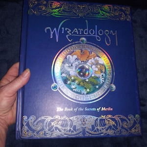 May include: A hardback book titled "Wizardology" with a blue cover and silver foil detailing. The cover features a circular design with illustrations and the text "The Book of the Secrets of Merlin". The book is held by a hand.