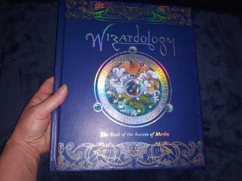 May include: A hardback book titled "Wizardology" with a blue cover and silver foil detailing. The cover features a circular design with illustrations and the text "The Book of the Secrets of Merlin". The book is held by a hand.