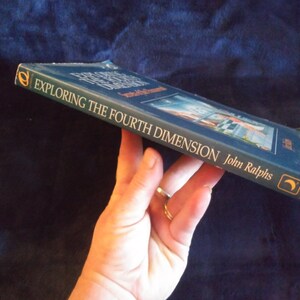 May include: A blue hardcover book titled "Exploring the Fourth Dimension" by John Ralphs. The book cover features a painting of a city skyline with a crescent moon in the upper right corner.