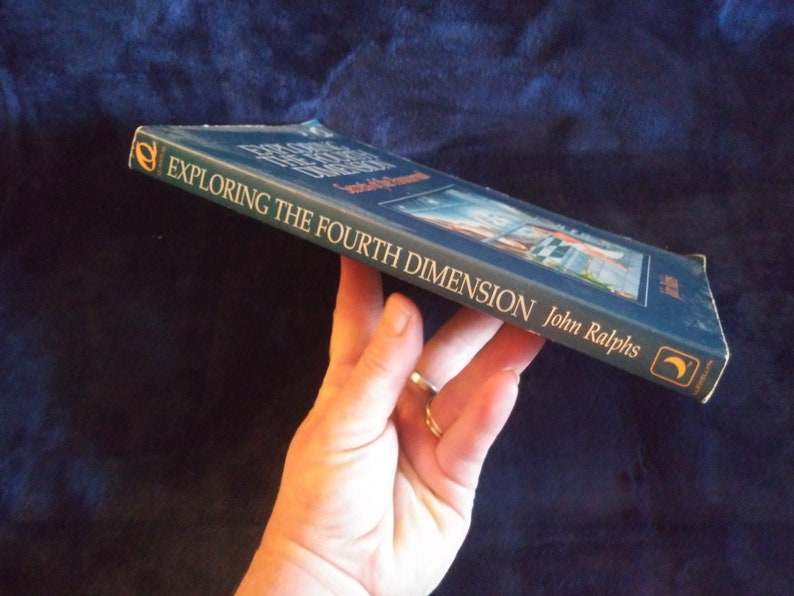 May include: A blue hardcover book titled "Exploring the Fourth Dimension" by John Ralphs. The book cover features a painting of a city skyline with a crescent moon in the upper right corner.