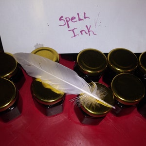 May include: A collection of small glass jars with gold lids filled with dark ink. A white feather with yellow accents rests on top of the jars. A piece of paper with the words "Spell Ink" written in purple ink is in the background.