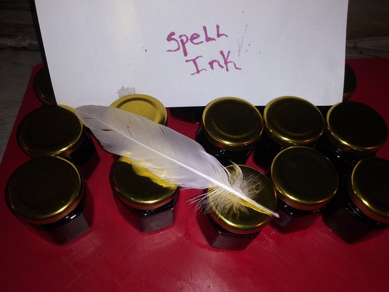 May include: A collection of small glass jars with gold lids filled with dark ink. A white feather with yellow accents rests on top of the jars. A piece of paper with the words "Spell Ink" written in purple ink is in the background.