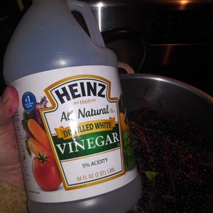 May include: A clear plastic bottle of Heinz All Natural Distilled White Vinegar, 64 fl oz (2 qt) 1.89L, held in front of a stainless steel pot. The label features a tomato, carrots, and an onion.