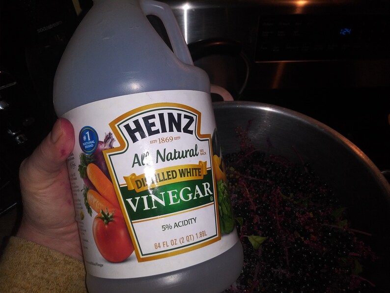 May include: A clear plastic bottle of Heinz All Natural Distilled White Vinegar, 64 fl oz (2 qt) 1.89L, held in front of a stainless steel pot. The label features a tomato, carrots, and an onion.