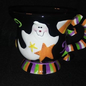 May include: Black ceramic Halloween mug featuring a smiling ghost with a yellow star and orange star. The handle is striped with orange, green, purple, and white. The base is striped with purple, green, and orange.