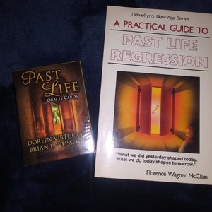 Puede incluir: Un libro titulado "A Practical Guide to Past Life Regression" de Florence Wagner McClain, parte de la serie New Age de Llewellyn. La portada del libro presenta una puerta con una luz brillante que brilla a trav&eacute;s. Junto al libro hay una baraja de cartas de or&aacute;culo titulada "Past Life Oracle Cards" de Doreen Virtue y Brian L. Weiss, M.D.
